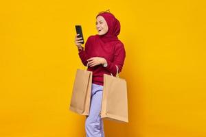 joyeuse jeune femme asiatique lisant un message sur un smartphone et montrant des sacs à provisions sur fond jaune photo