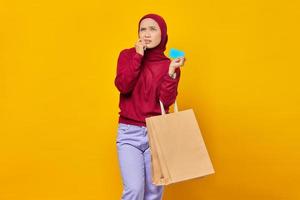 Jeune femme asiatique confuse mordant la carte de crédit et tenant le sac à provisions sur fond jaune photo