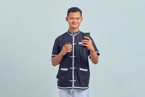 Portrait d'un jeune homme asiatique souriant portant un uniforme de karaté utilisant un smartphone et tenant un café sur fond gris photo