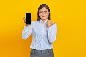 photo d'une femme d'affaires asiatique souriante montrant un écran de smartphone vierge et pointant vers un espace vide avec le pouce