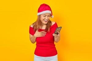 Jeune femme asiatique excitée en chapeau de père Noël tenant un téléphone portable avec les mains sur la poitrine sur fond jaune photo