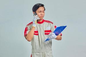 portrait de jeune mécanicien asiatique parlant au téléphone et tenant le presse-papiers avec une expression confuse isolée sur fond gris photo