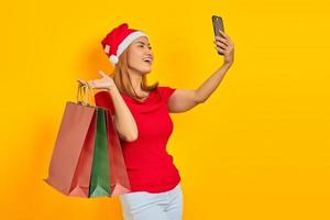 Happy young asian woman in santa claus hat holding shopping bags et prenant selfie sur mobile sur fond jaune photo