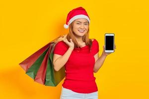 Souriante jeune femme asiatique en chapeau de père Noël montrant un téléphone portable à écran blanc et tenant des sacs à provisions isolés sur fond jaune