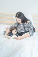 famille aimante heureuse jeune mère asiatique lisant un livre drôle avec sa fille mignonne dans la chambre à coucher. conception de photo pour la famille, les enfants et le concept de personnes heureuses