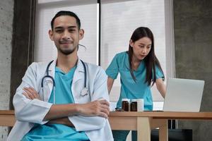 portrait d'un jeune médecin d'origine asiatique en uniforme avec stéthoscope. sourire et regarder la caméra dans une clinique hospitalière, partenaire féminin travaillant derrière, professionnel de la médication à deux personnes.