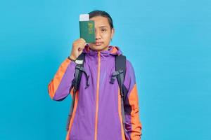 Portrait d'un jeune homme asiatique portant une veste violette couvrant un œil avec un livre de passeport isolé sur fond bleu photo