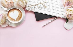 bureau pour femmes vue de dessus à plat avec des fleurs. espace de travail féminin avec ordinateur portable, pivoines de fleurs, accessoires, ordinateur portable, lunettes, tasse de café sur fond rose. photo