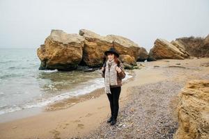 femme dans un chapeau et une écharpe avec un sac à dos sur la côte sur fond de rochers contre une mer magnifique photo