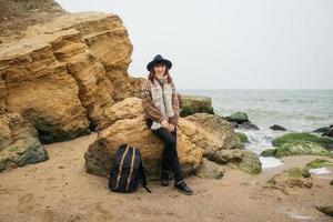femme dans un chapeau et une écharpe avec un sac à dos sur la côte sur fond de rochers contre une mer magnifique photo