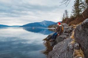 jeune homme avec un sac à dos jaune portant un chapeau rouge assis sur le rivage sur fond de montagne et de lac. espace pour votre message texte ou contenu promotionnel. concept de mode de vie de voyage. photo