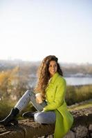 jolie jeune femme profitant du soleil d'automne assis au bord de la rivière et buvant du café à emporter photo