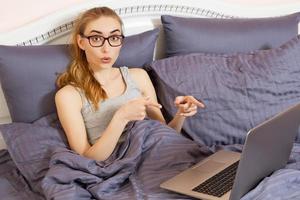 belle femme surprise dans des verres pointant sur son ordinateur portable allongé sur son lit. fille travaillant sur ordinateur portable à la maison chambre photo