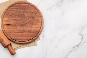 plateau de pizza en bois vide avec serviette et rouleau à pâtisserie mis en place sur une table de cuisine en pierre de marbre. planche à pizza et nappe sur fond de marbre blanc. photo