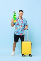 souriant bel homme de tourisme asiatique voyageant avec un pistolet à eau et des bagages pendant le studio du festival de songkran tourné sur fond bleu photo