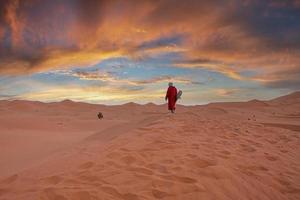 L'homme en vêtements traditionnels avec sandboard marchant sur les dunes de sable au crépuscule photo