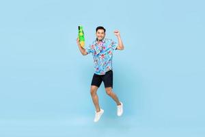 beau jeune homme asiatique souriant jouant avec un pistolet à eau et sautant en studio sur fond bleu pour le festival de songkran en thaïlande et en asie du sud-est photo