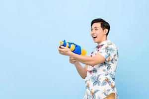 souriant bel homme asiatique jouant avec un pistolet à eau isolé sur fond bleu studio pour le festival de songkran en thaïlande et en asie du sud-est photo