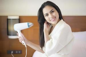 Souriante jeune femme portant un peignoir blanc séchant ses cheveux avec un sèche-cheveux après une douche dans la chambre