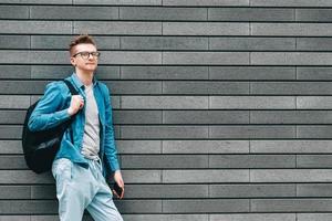 homme en chemise, jeans et lunettes portant un sac à dos et tenant un smartphone en se tenant debout sur un fond de mur gris