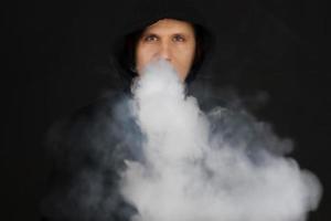 l'homme fume une cigarette électronique sur le fond sombre, les hommes en sweat à capuche vapotent et libèrent un nuage de vapeur. mec avec vapotage sur fond noir photo