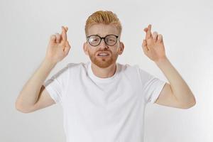 croisons les doigts, faites un concept de souhait. jeune homme rousse avec un doigt croisé de barbe rouge pour la bonne chance. signe de la main, geste pour que les rêves deviennent réalité. homme en t-shirt blanc modèle vierge. maquette, espace de copie photo