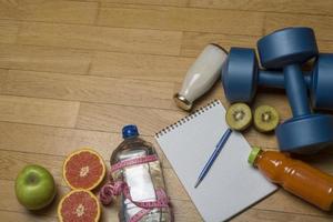 entraînement, exercice, gaieté et santé - deux haltères en plastique, un cahier, de l'eau minérale avec du jus, des fruits et un stylo sur le parquet. photo