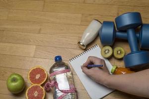 entraînement, exercice, gaieté et santé - deux haltères en plastique, eau minérale avec jus, fruits et main écrit dans un cahier sur le parquet. photo