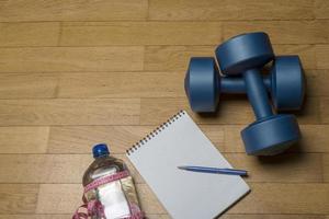 entraînement, exercice, gaieté et santé - deux haltères en plastique, un cahier, de l'eau minérale et un stylo sur le parquet. photo