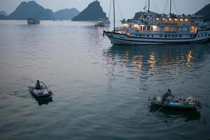 baie d'halong, vietnam - 08242015 - petits bateaux s'approchant des navires de croisière pour vendre leurs marchandises aux touristes. fin d'après-midi, lumières allumées.