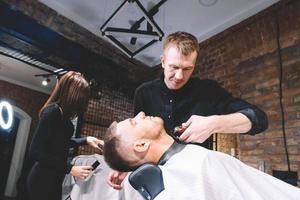 le jeune coiffeur masculin fait un client de barbe de coupe de cheveux avec un rasoir droit vintage. processus de rasage des barbes dans le salon de coiffure photo