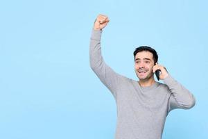portrait d'un beau jeune homme de race blanche souriant avec le poing levé parlant au téléphone en entendant de bonnes nouvelles dans un studio isolé sur fond bleu