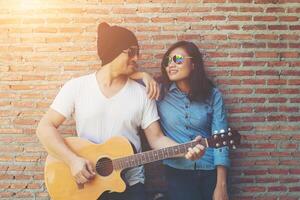 un couple hipster se serre dans ses bras, regarde au loin et sourit tout en se tenant debout en plein air jouant de la guitare contre un mur de briques, les rencontres ont passé du bon temps ensemble. photo