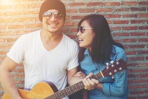 un couple hipster se serre dans ses bras, regarde au loin et sourit tout en se tenant debout en plein air jouant de la guitare contre un mur de briques, les rencontres ont passé du bon temps ensemble. photo