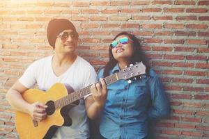 un couple hipster se serre dans ses bras, regarde au loin et sourit tout en se tenant debout en plein air jouant de la guitare contre un mur de briques, les rencontres ont passé du bon temps ensemble. photo