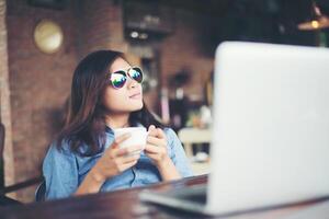 belle jeune femme hipster assise dans un café, tenant une tasse de café, détendez-vous et jouez avec son ordinateur portable, regardant ailleurs, heureuse et amusante. notion de style de vie. photo