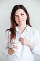 portrait d'une belle femme dans une chemise blanche avec une tasse en céramique dans ses mains sur un fond blanc. femme buvant de l'eau café thé de tasse tasse blanche photo