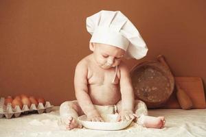 petit garçon boulanger dans une toque de chef assis sur la table jouant avec de la farine sur un fond marron avec un rouleau à pâtisserie en bois, un tamis rustique rond et des œufs. copie, espace vide pour le texte photo