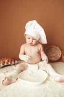 petit garçon boulanger dans une toque de chef assis sur la table jouant avec de la farine sur un fond marron avec un rouleau à pâtisserie en bois, un tamis rustique rond et des œufs. copie, espace vide pour le texte photo