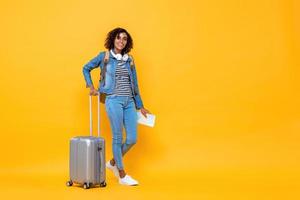 portrait de voyage pleine longueur d'une jeune femme afro-américaine souriante routard debout et tenant des bagages sur fond de studio jaune photo