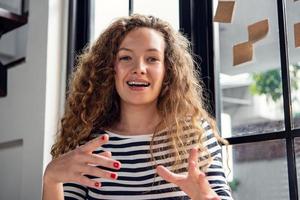 femme caucasienne joyeuse regardant la caméra en souriant et en parlant tout en faisant une conférence téléphonique vidéo à la maison, concept de réunion à distance photo