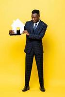 jeune homme d'affaires africain en tant qu'agent immobilier tenant un modèle de découpe de maison en arrière-plan de studio isolé jaune photo