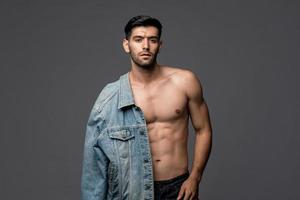 Jeune bel homme à moitié torse nu sexy avec une veste en jean posant en fond de studio isolé gris clair photo