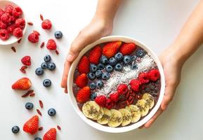 mains de femme tenant un bol blanc de smoothies de superaliments aux baies d'açai avec graines de chia, fraise, baie de goji, noix de coco, myrtille, garnitures de framboise et pépites de chocolat sur fond blanc photo