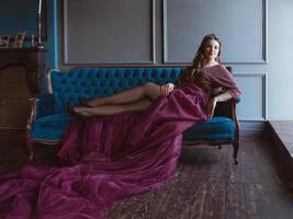 portrait de la belle jeune princesse jolie jolie femme en longue robe et couronne de reine violette, avec de longs cheveux et maquillage assis sur le canapé en velours bleu à l'intérieur dans un intérieur minimaliste photo