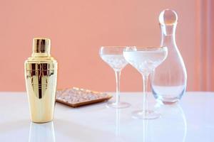 shaker doré, carafe en verre, verres élégants avec cocktail et glace sur fond beige. alcool, fête, hôtel, concept de bar photo