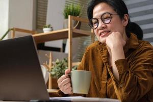 une femme asiatique boit une tasse de café et regarde la diffusion en direct sur un ordinateur portable lorsqu'elle travaille à la maison photo