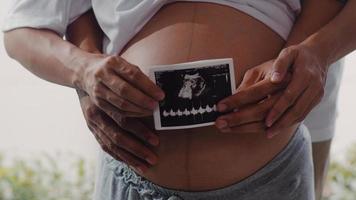 jeune couple enceinte asiatique montre et regarde un bébé photo échographique dans le ventre. maman et papa se sentant heureux souriant paisible tout en prenant soin de l'enfant allongé près de la fenêtre dans le salon à la maison concept.