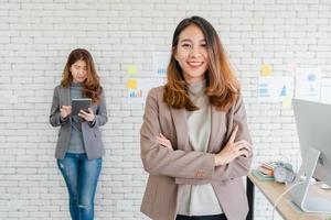 deux jeunes femmes d'affaires créatives asiatiques tenant une tasse de café, travaillant sur un ordinateur portable et discutant du travail tout en travaillant au bureau. affaires occasionnelles, travail indépendant au café, concept de réunion sociale. photo