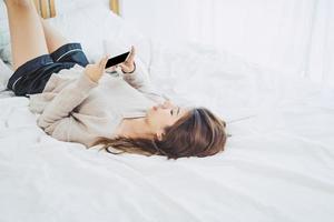 les femmes asiatiques heureuses utilisent un téléphone intelligent avec un écran vide noir sur le lit le matin. femme asiatique au lit vérifiant les applications sociales avec smartphone. femme souriante surfant sur le net avec un téléphone portable à la maison. photo